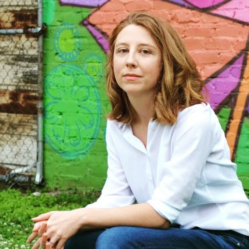 Individual with medium length hair sits with elbows on thighs in front of a multicolored wall.