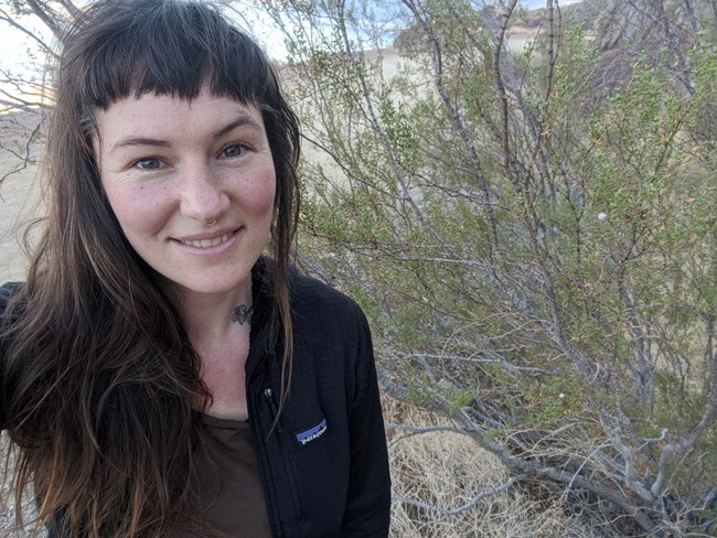 Woman smiling, standing next to desert shrubland vegetation.