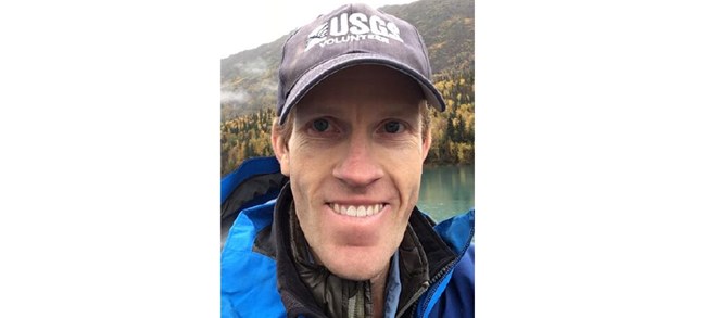 Carl Legleiter Legleiter smiling outside in a USGS ball cap in front of a scenic lake.