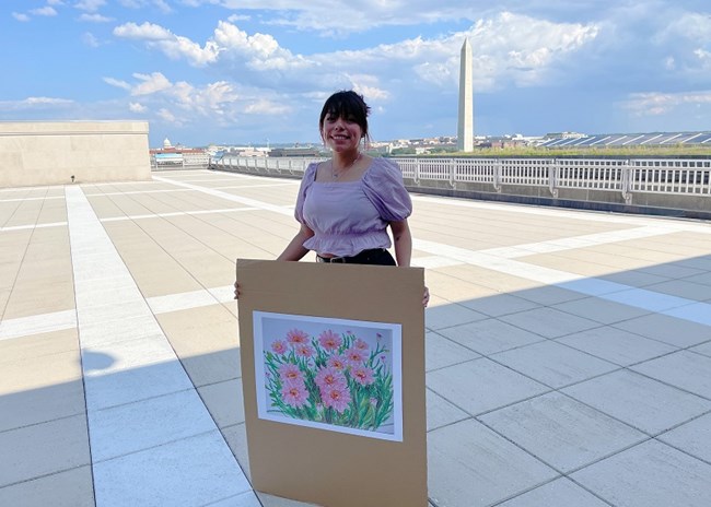 person holding a picture of an oil painting of pink flowers, with the Washington Monument in the background.