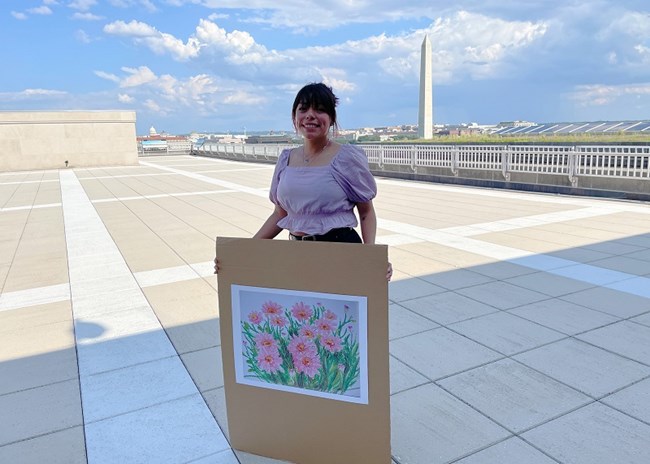 Leslie Garcia person holding a picture of an oil painting of pink flowers, with the Washington Monument in the background.