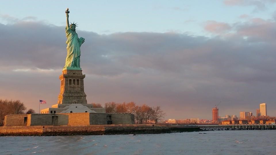 A sunset photo of Liberty Island, with the Statue of Liberty standing tall holding aloft her torch