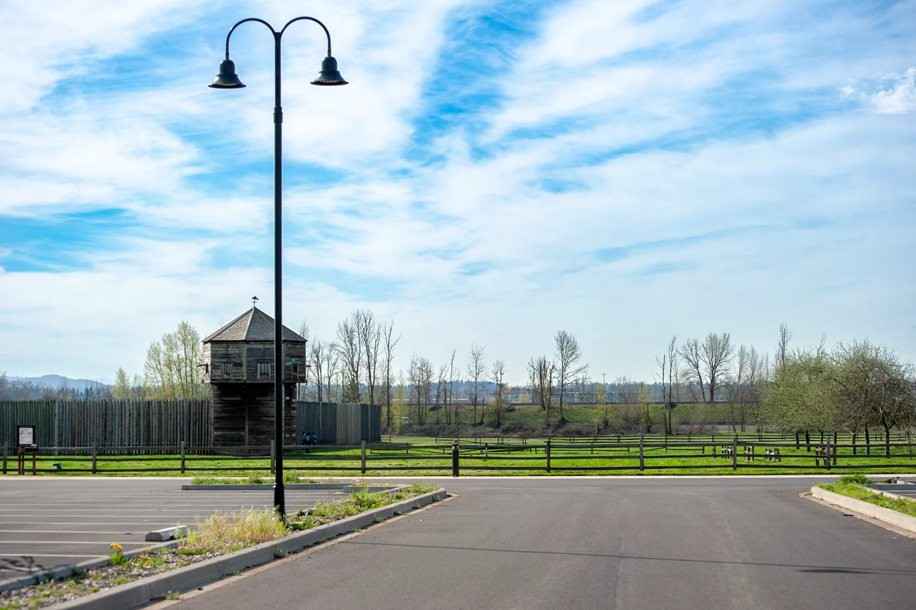 Light Pole_Bastion A black lightpost, tall and with two hooks each holding a light, is shown in an empty parking lot. In the background is a wooden bastion, an octagonal watchtower in the corner of a wooden palisade.