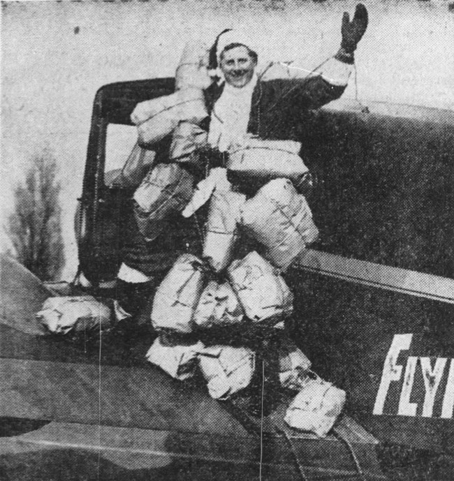 A man in a santa suit carries presents as he stands on the wings of a plane