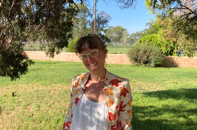 A women with light hair pulled back, stands underneath a tree.