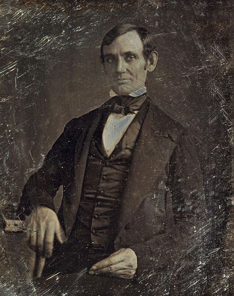 A black and white photo of 38-year-old Abraham Lincoln. Lincoln is seated, looking straight into the camera. He is clean-shaven, with his hair combed close to his head. He wears a black suit and bow tie.