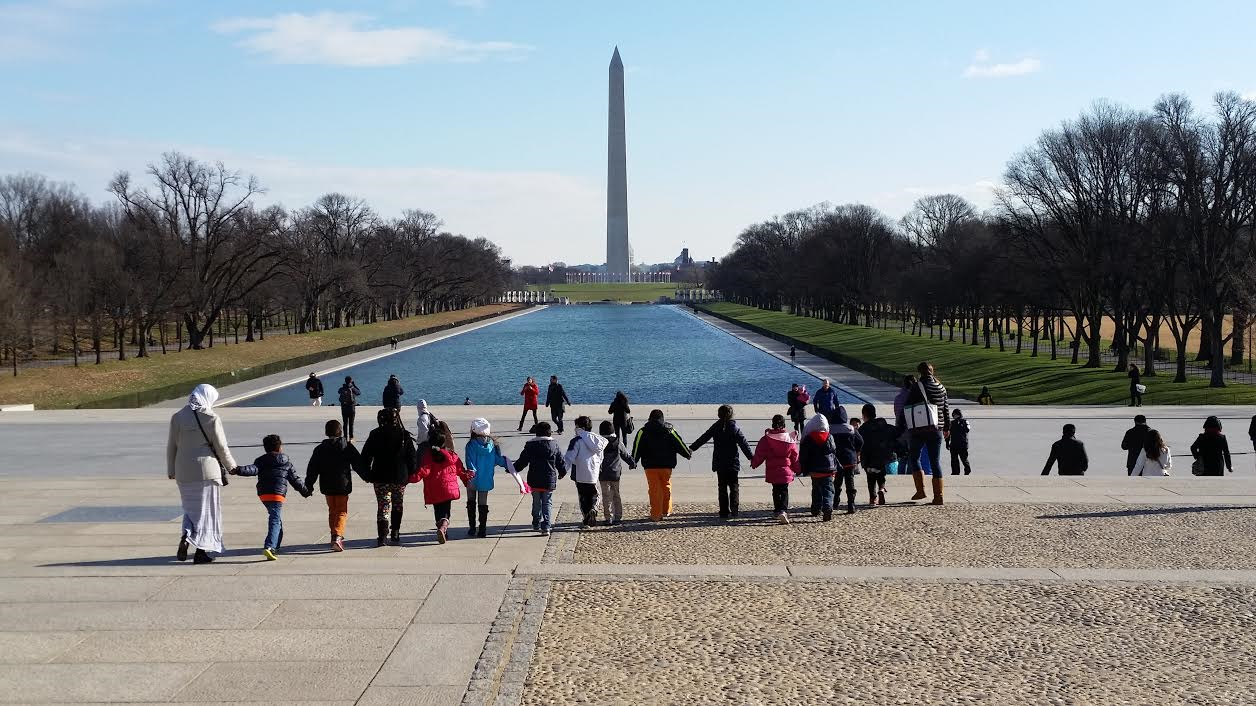 Lincoln Plaza WAMO Students on a field trip hold hands while walking near the Reflecting Pool with Washington Monument in the distance