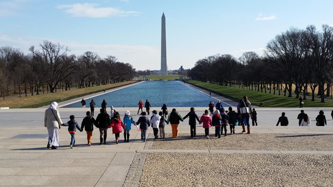 Students on a field trip hold hands while walking near the Reflecting Pool with Washington Monument in the distance