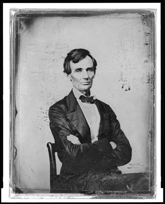 Abraham Lincoln, beardless and sitting in a chair, arms crossed.