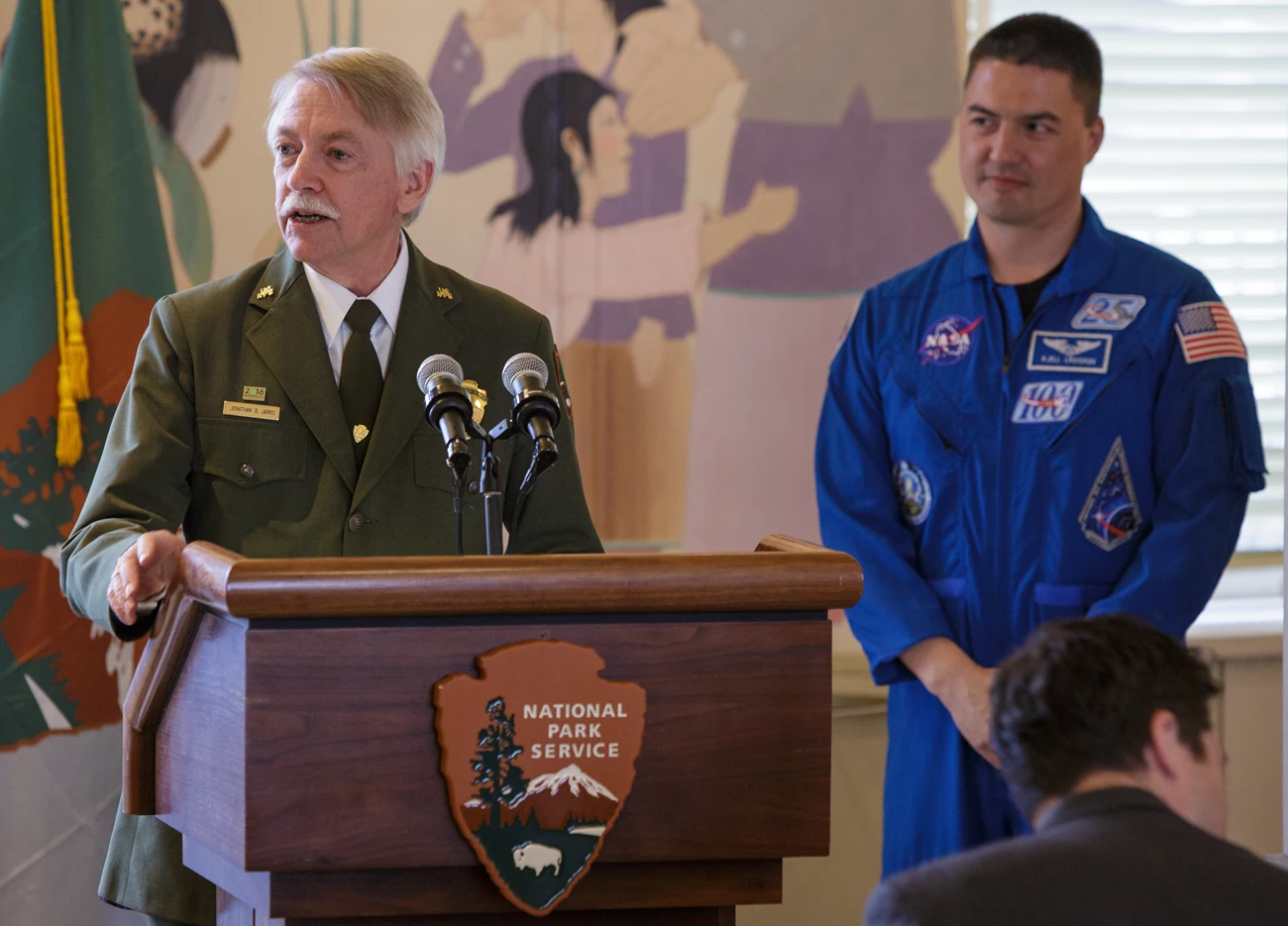 Jarvis and Lindgren Director Jarvis in his NPS uniforms stands at a podium as Astronaut Kjell Lindgren, in a blue flight suit, looks on.