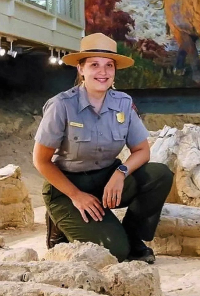 A person wearing a park ranger uniform and hat is kneeling next to large fossilized bones with a mural of mammoths in the background.