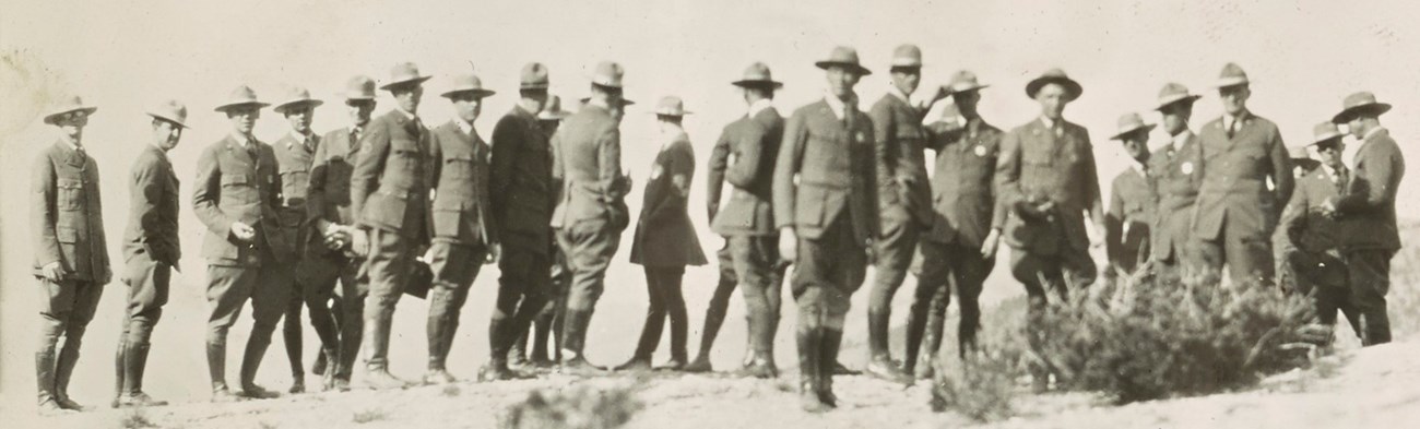 A large group of rangers stand in a line wearing their full ranger uniforms.  Some face away and some look over their shoulders.