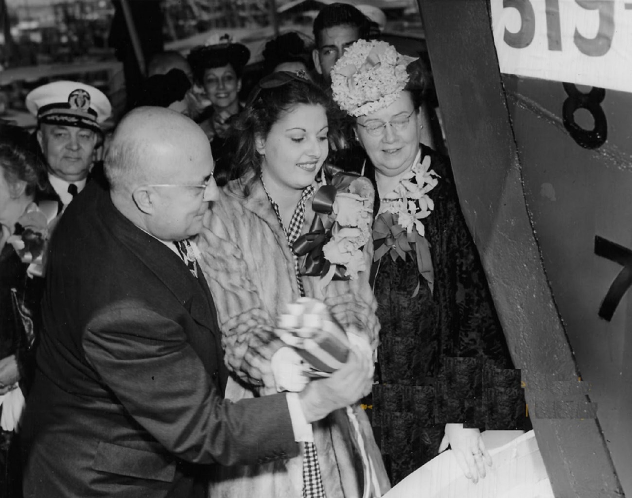 two women and one man hold champagne bottle next to front of large ship