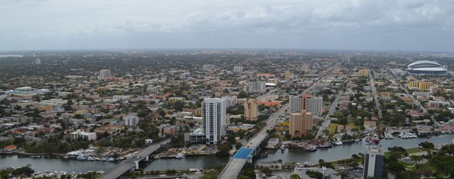 Aerial view of Little Havana neighborhood in Miami, showing Marlins Park at the right, the Coral Gables skyline in the background, and Coconut Grove at the left. Two bridges over a river are visible in the foreground.