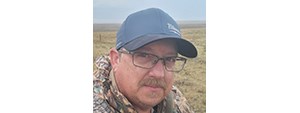 Man with a cap, glasses, and mustache, outside in a vast grassland.
