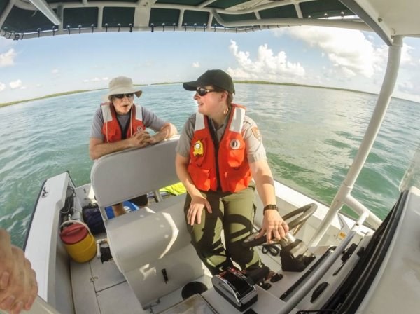 Liz drives a boat with a passenger sitting behind her. Both people are wearing life vests.