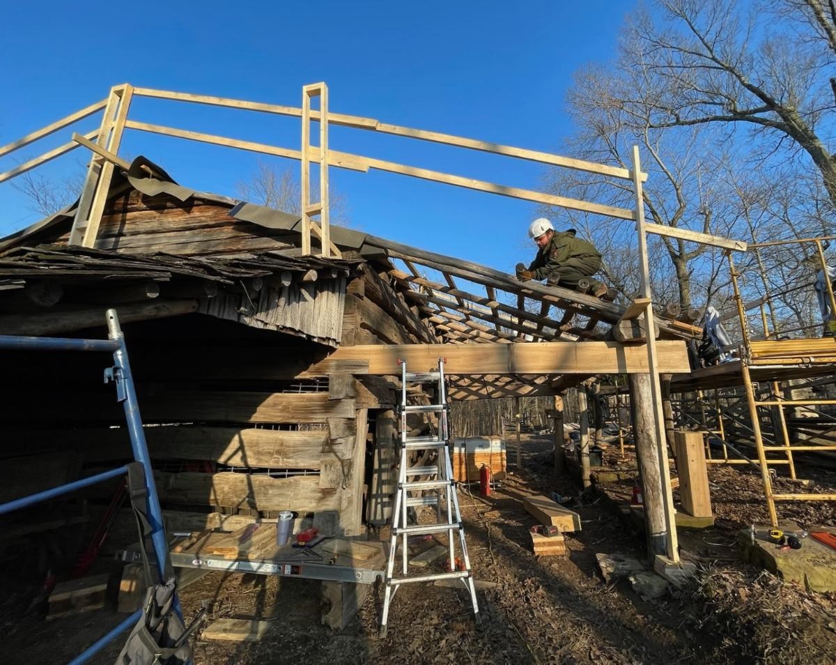 Worker wearing white helmet and goggles safety gear while perched on his hands and knees on log beams on a barn.