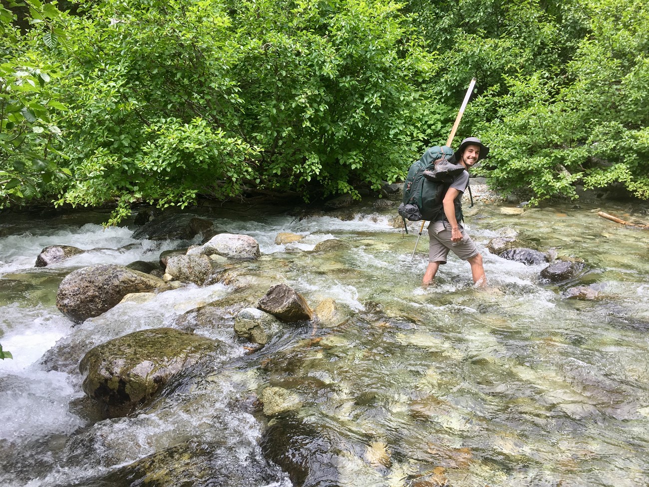 A person with a large backpack wading through a rushing, rocky river and smiling at the camera.