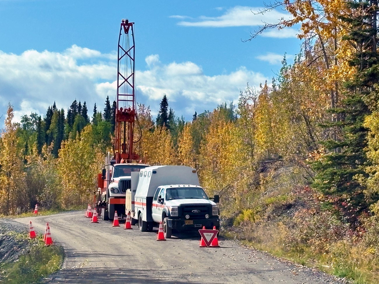 A large, white truck with a tower, on a dirt road lined with evergreen trees.