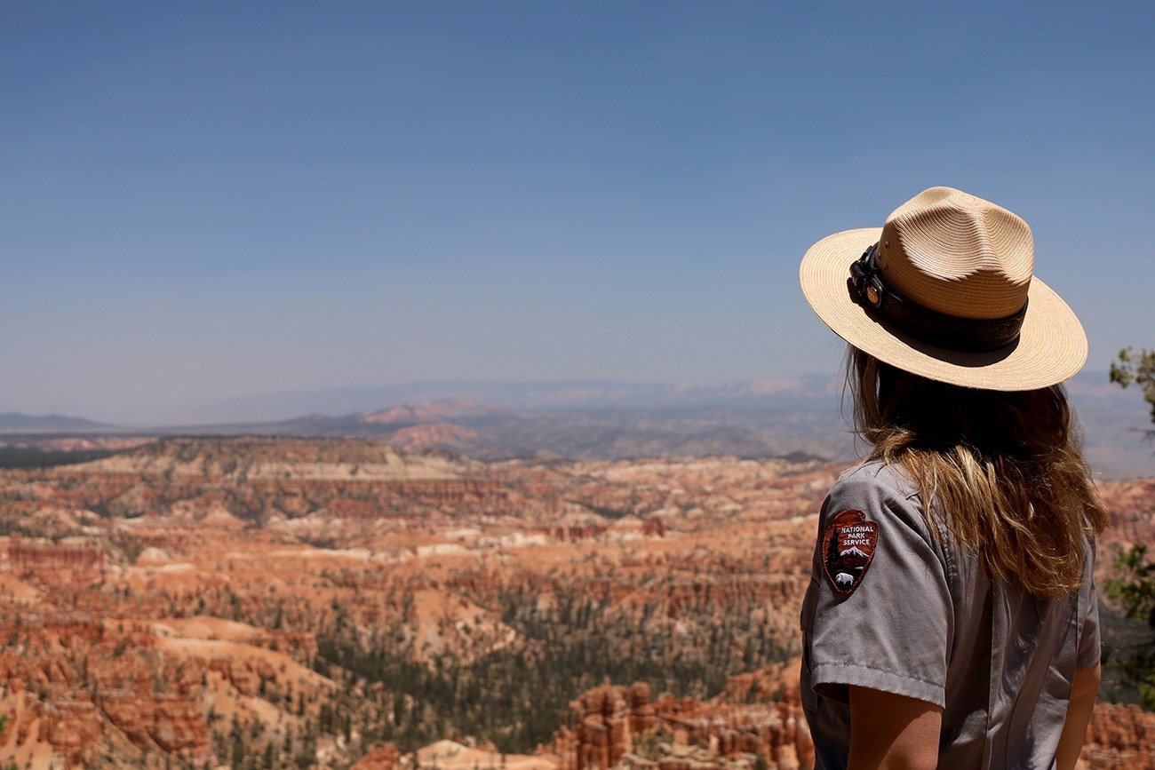 A woman with long hair and in an NPS uniform and hat stands with her back to the camera as she looks at a distant landscape filled with red rock formations.