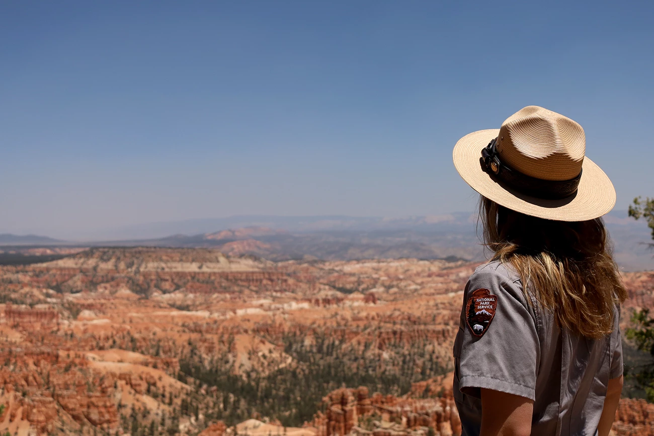 Looking out over Bryce Canyon A woman with long hair and in an NPS uniform and hat stands with her back to the camera as she looks at a distant landscape filled with red rock formations.