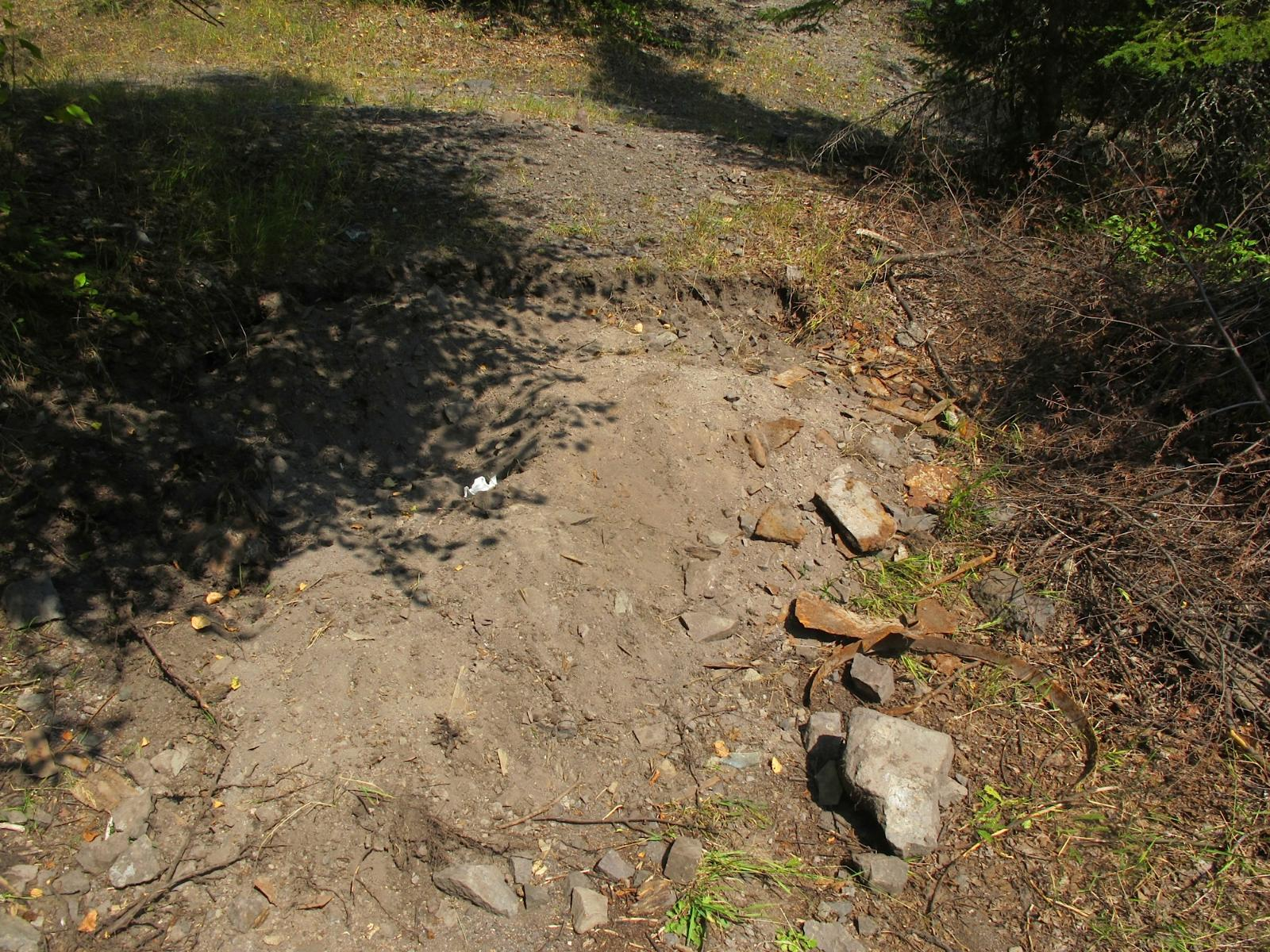 A man-made hole with a mound of brownish gray rocks next to it with a scattering of rusty metal pieces.