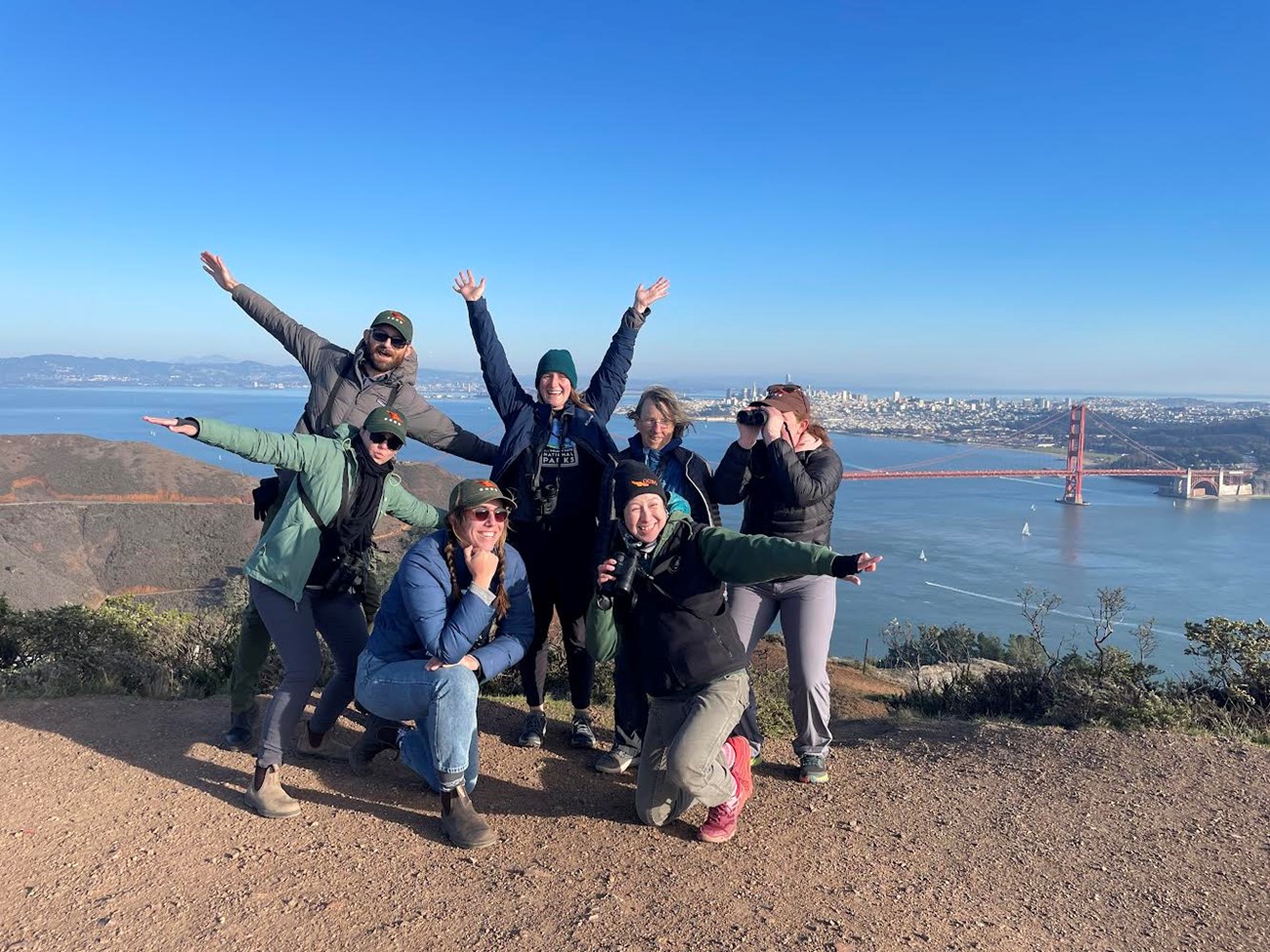 7 Golden Gate Raptor Observatory volunteers with binoculars posing in front of the Golden Gate Bridge on a hill on a clear day with the cityscape behind them.