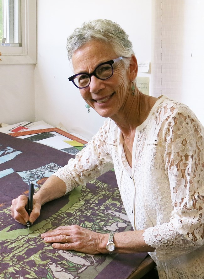 Smiling woman with short white hair, dark glasses, and a lace blouse leans over artwork woth a pen in her hand