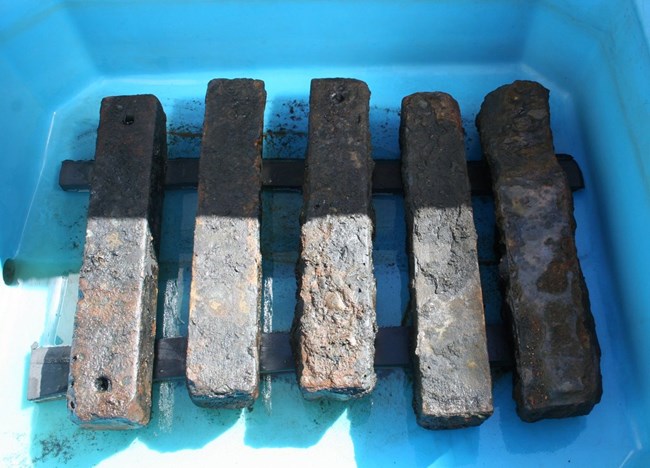 Five rusted metal bars side by side from left to right on a blue background. They are connected by two perpendicular thinner metal bands, like a railroad.