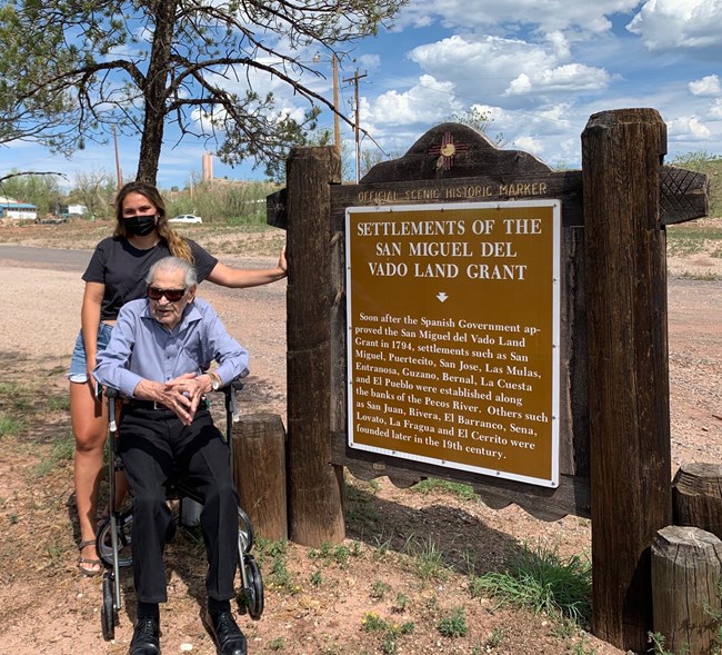 A man in a wheelchair and a young lady behind him, next to a large brown sign.