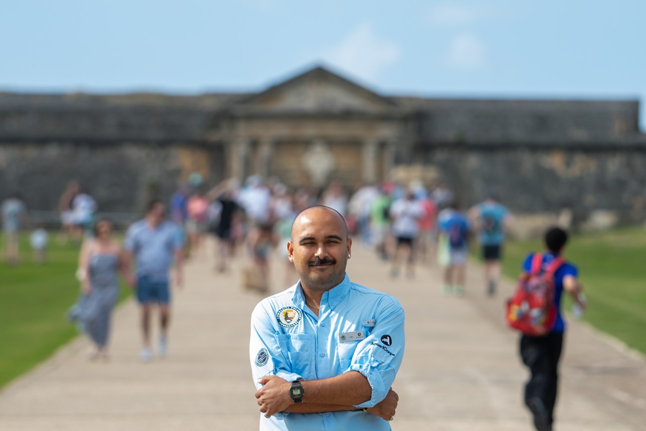 A young man smiles in front of Castillo San Felipe del Morro, with blurred people walking in the background.
