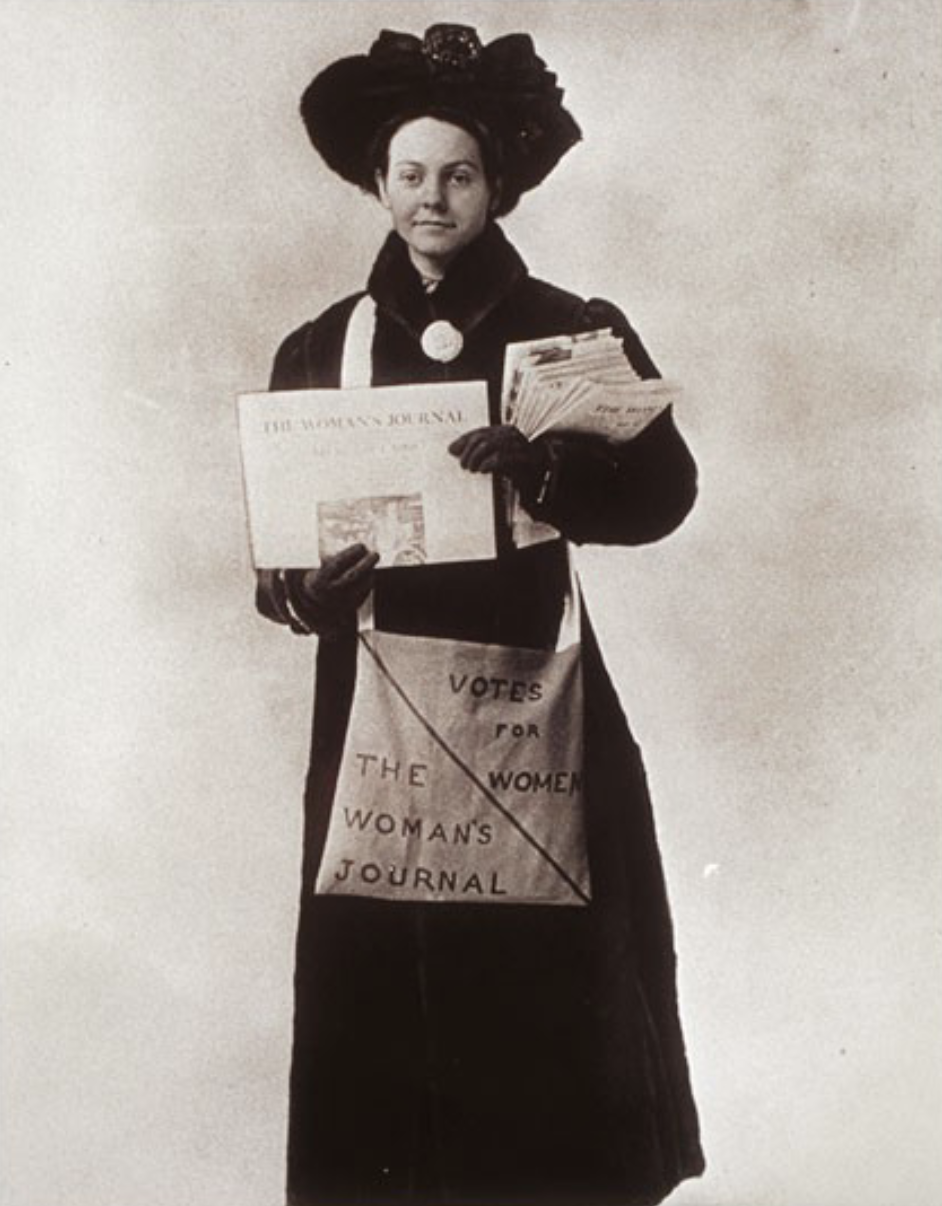a woman wearing a long winter coat, hat, and gloves, holding copies of the Woman's Journal and carrying a canvas bag that says "Votes for Women/The Woman's Journal"