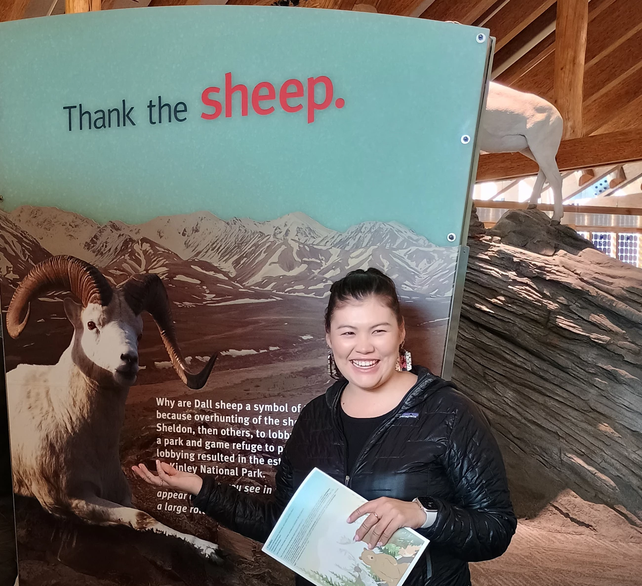 Alaska Region Tribal Liaison Lydia Agnus Agnus smiling and gesturing toward a visitor center display featuring a photo of a Dall sheep in the mountains with text that reads "Thank the sheep."