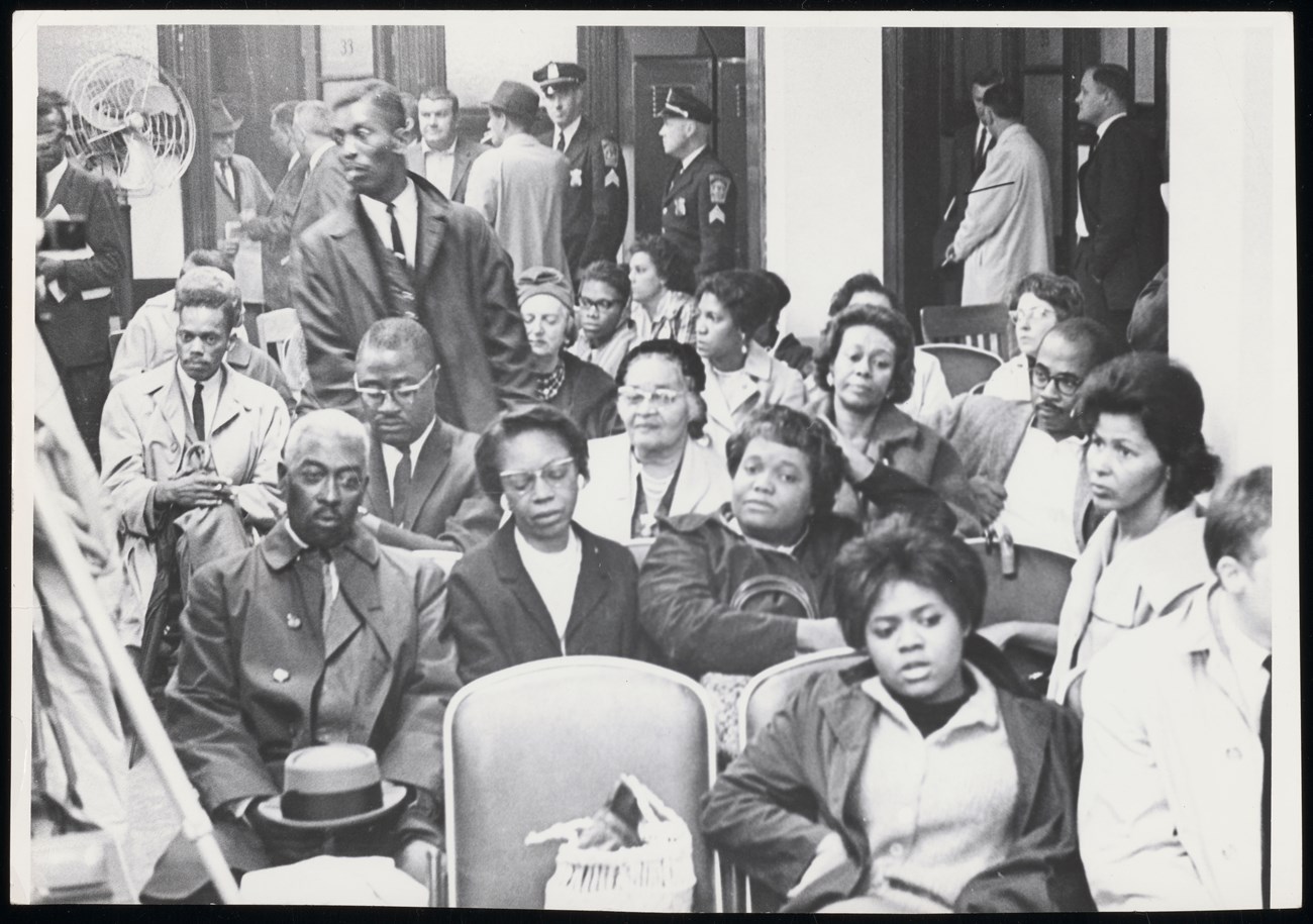 M214_1015935973_0024_0011_A Black and white image of middle aged Black folks sitting in chairs in an auditorium