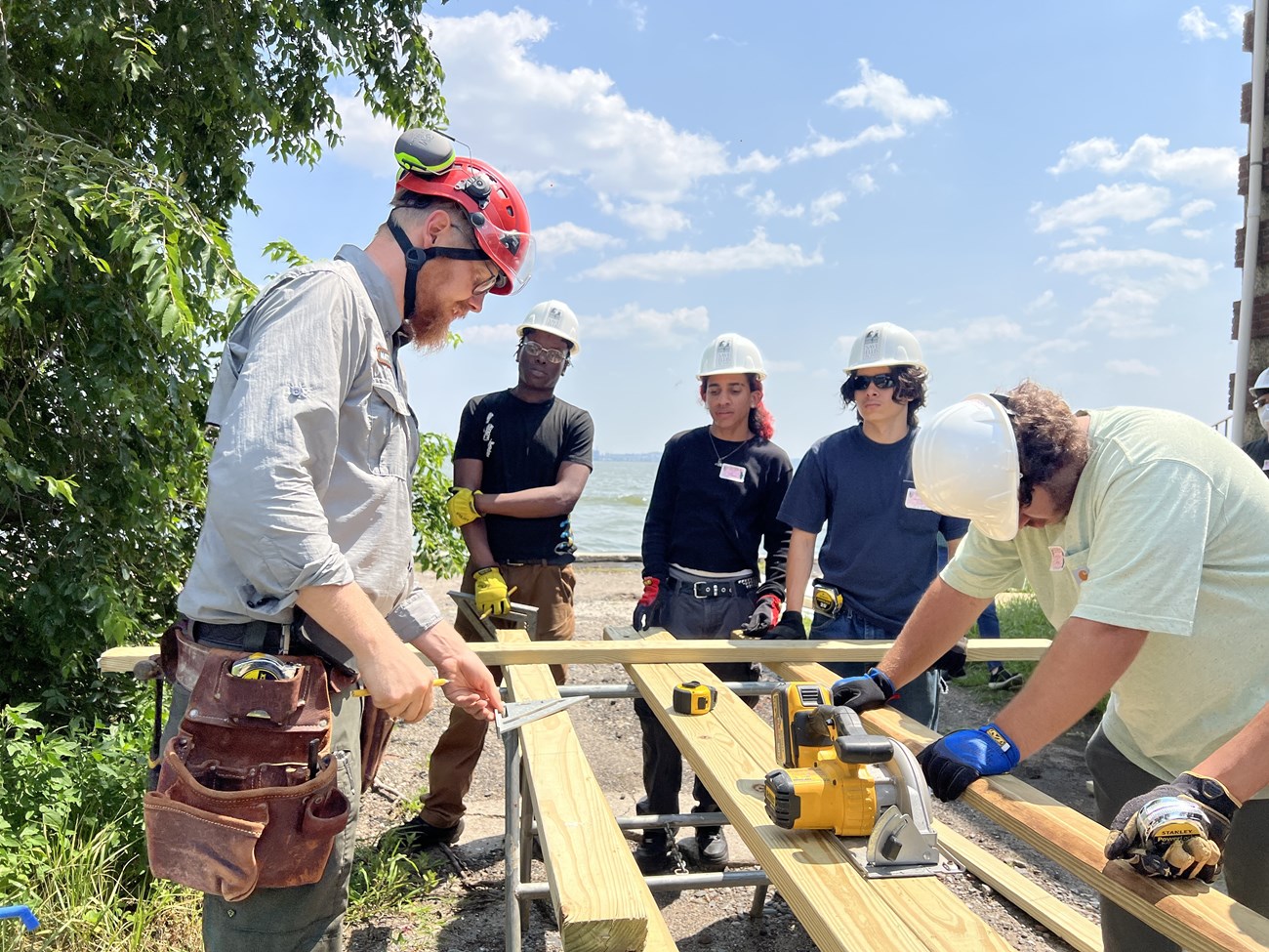 lt= A group of people stand outside wearing hard hats and surround a circular saw