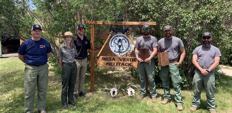Forrest Rackham and Tyrel Matthews were awarded the 2021 National Park Service Aviator of the Year Award. Five men and one woman stand surrounding the Mesa Verde Helitack sign; two men hold awards.