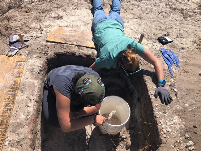 One figure crouches in a square archaeology dig while another reaches down into the hole