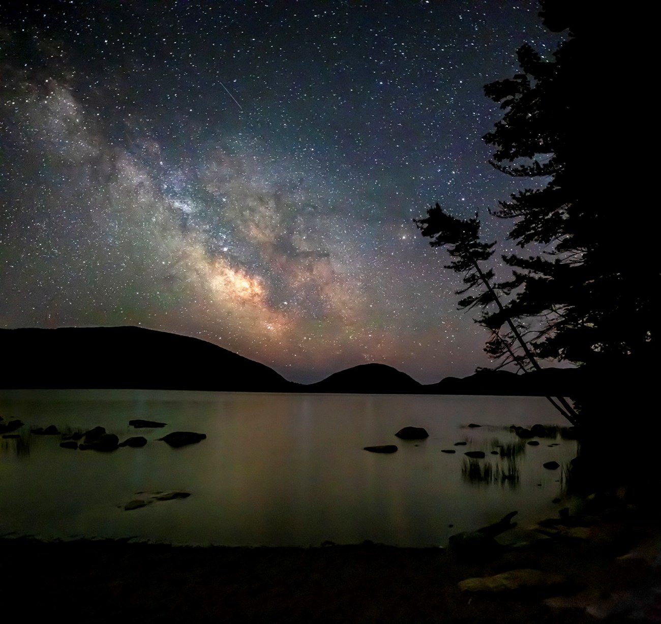 Night sky image with Milky Way in distance, a silhouette of mountains and lake reflections in middle distance, and silhouette of a leaning tree in foreground Night sky image with Milky Way in distance, a silhouette of mountains and lake reflections in middle distance, and silhouette of a leaning tree in foreground