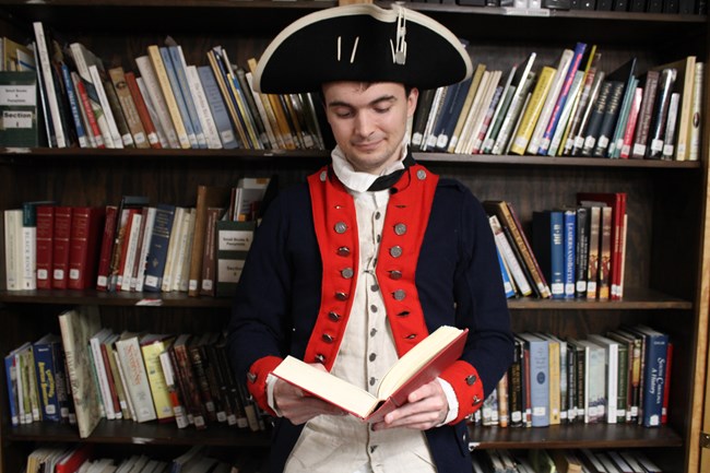 Reenactor in 18th-century military uniform reads book.