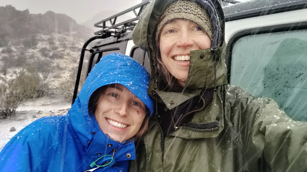 Desert Springs Monitoring Crew Selfie of two women wearing hooded raincoats smiling at camera in snowfall.
