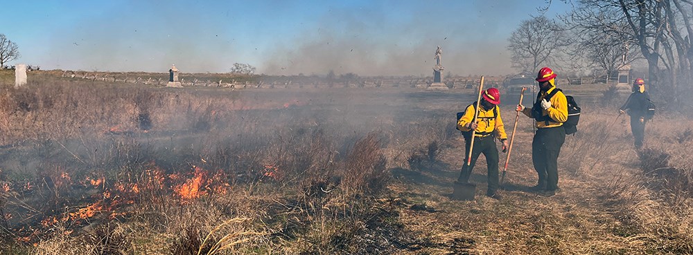 Wildland firefighters monitor a prescribed fire at Monocacy National Battlefield. Firefighters use handtools near a fireline with battlefield monuments in the distance.