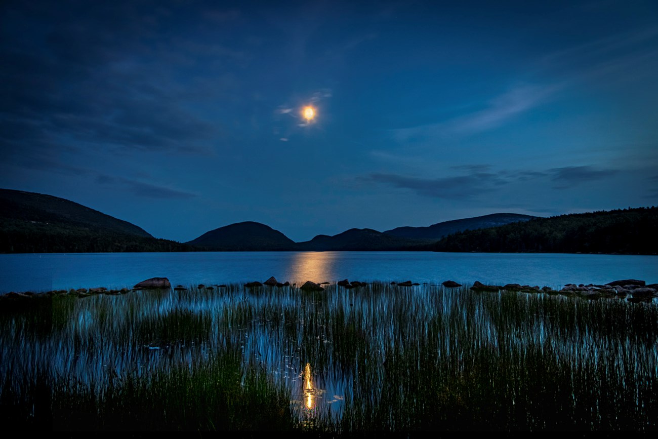 Night photo with dark blue sky, silhouette of mountains in distance, and moon hanging in center with reflections in water below Night photo with dark blue sky, silhouette of mountains in distance, and moon hanging in center with reflections in water below