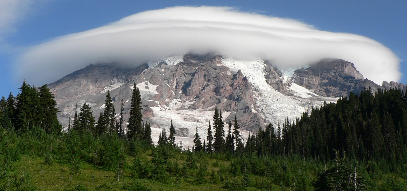 Large cloud circling a very tall mountain with an alpine forest at its base