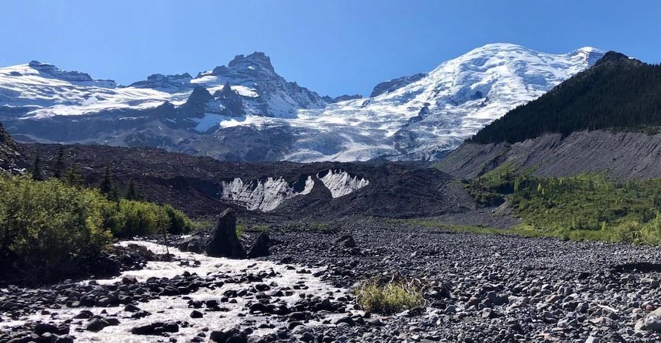 Emmons Glacier Terminus A glacier descends from a mountain peak ending in a rock-covered terminus with a river emerging from the ice into a rocky plain.