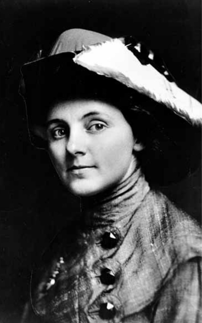 Young woman in large hat and high-necked shirt with large button smiles at camera