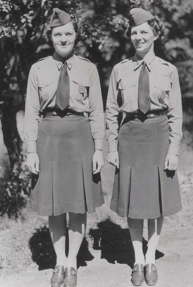 Two women stand posing to show off their NPS uniforms consisting of shirt, shirt, tie, badge, and cap.