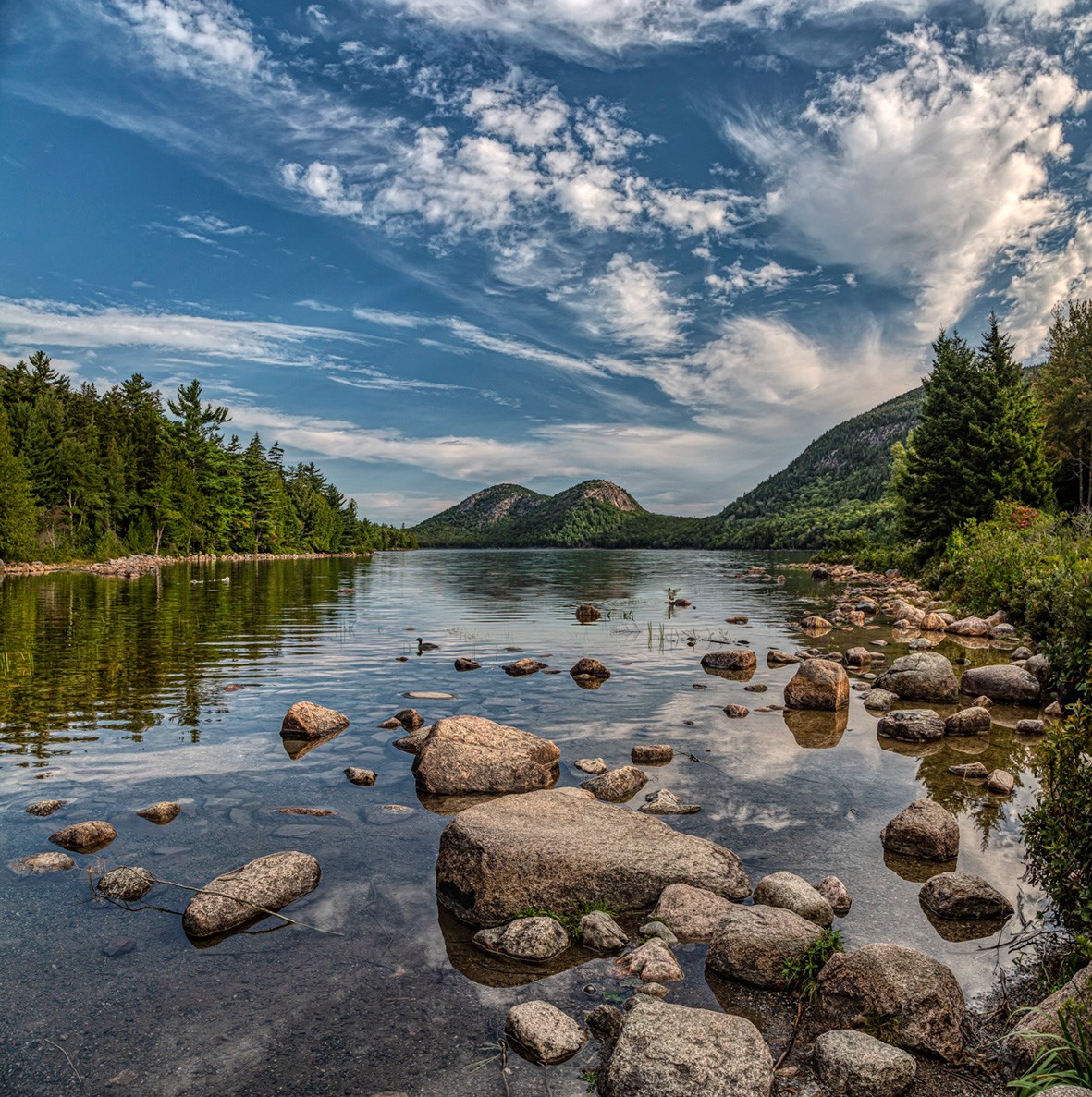 Landscape photo of a clear pond with pink boulders in the foreground, wooded hills along the shoreline and mountains and billowing clouds in the distance.