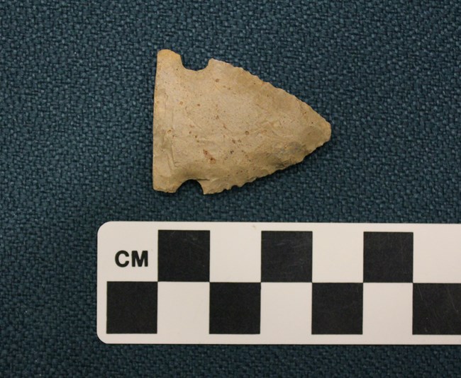 A beige triangular arrowhead with knapped edges, next to a black-and-white measuring tape; it is about 3 cm long.