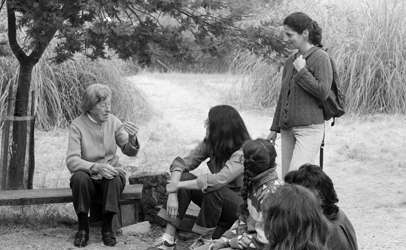 Older woman in sweater sits under tree, talking to young students who surround her