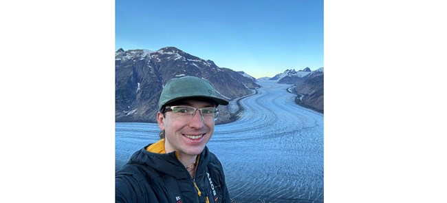 selfie of man with glasses in a cap and jacket in front of a glacier
